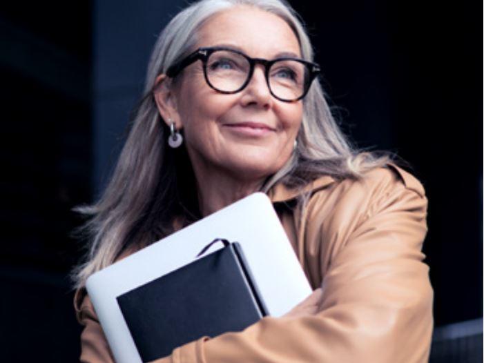 A picture of an older woman with glasses smiling and carrying a laptop.
