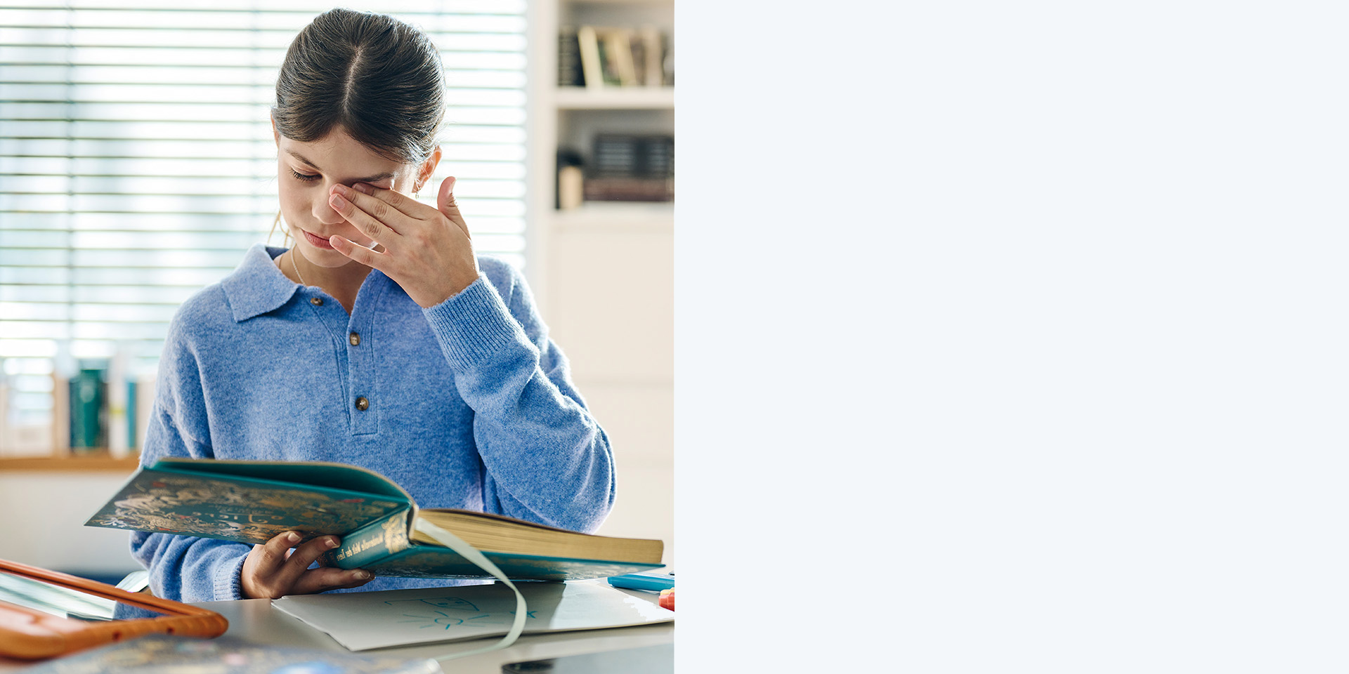 Young brown-haired girl without glasses rubbing her eyes while reading a book.