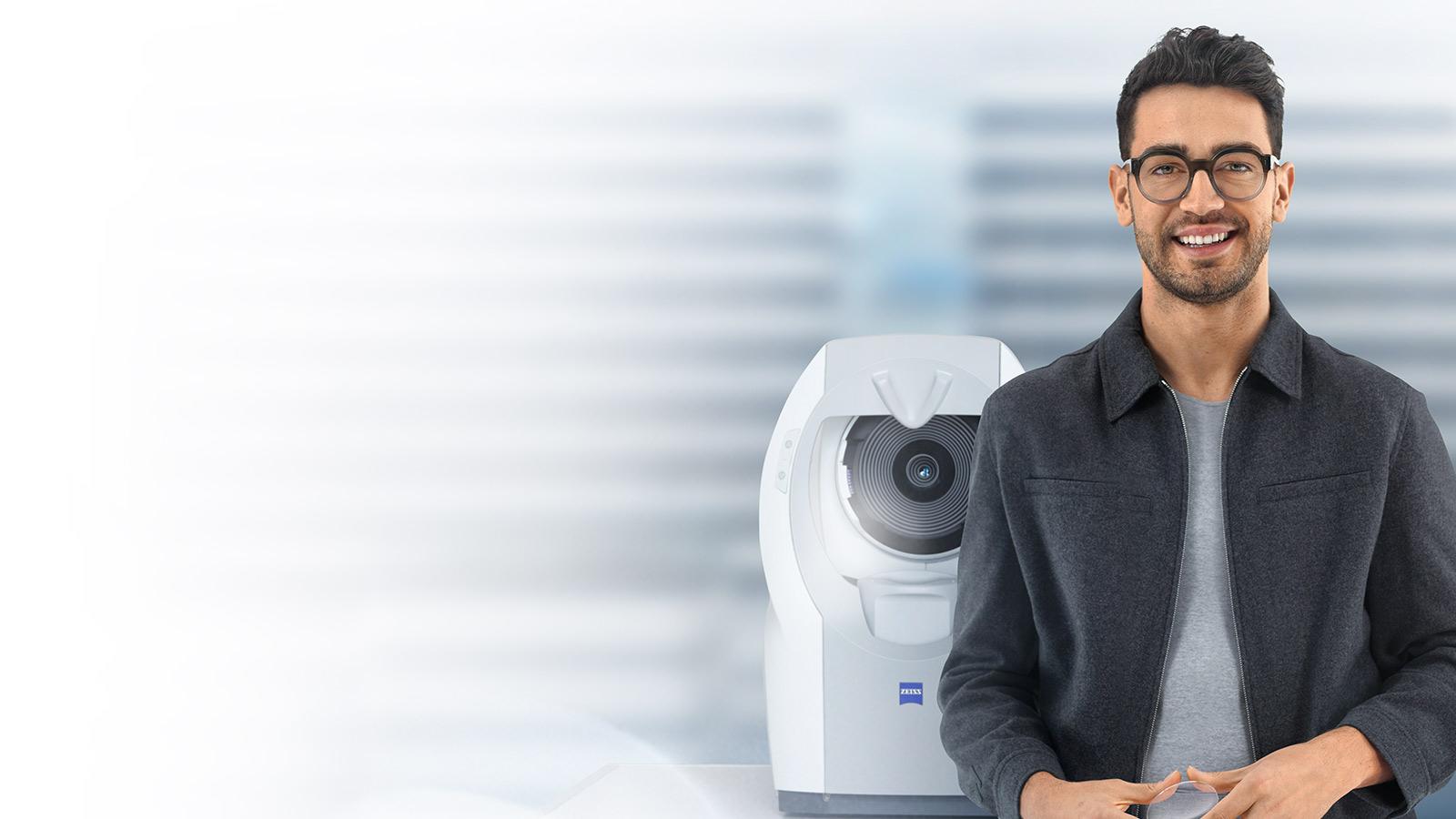 Young man with short brown hair smiling in camera wearing ZEISS lenses. In the background there is a ZEISS i.Profiler plus.