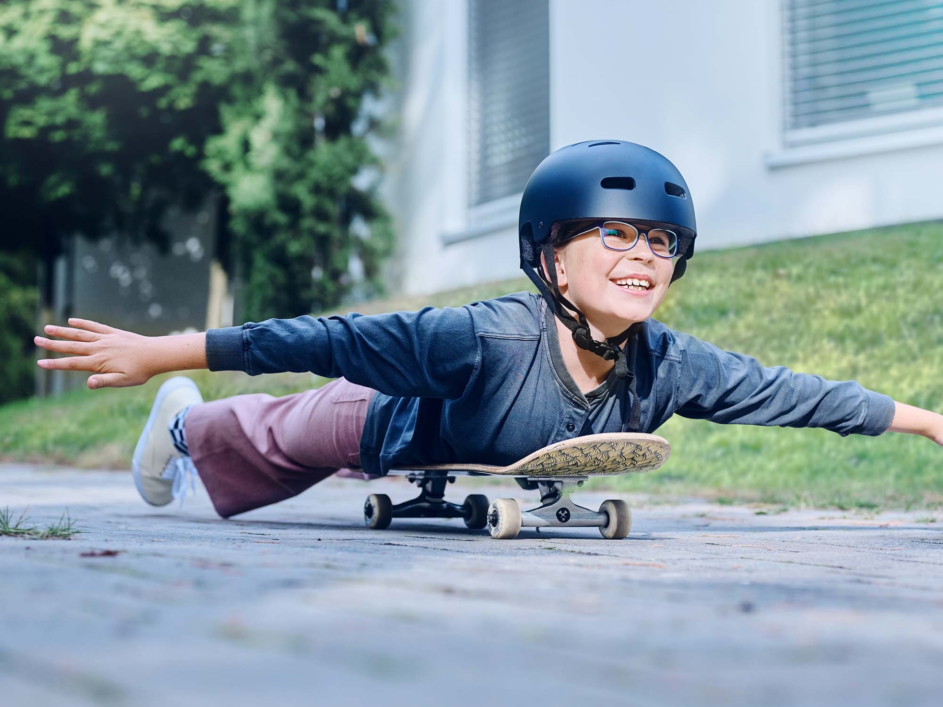 A girls with a helmet and glasses rolls lying on a skateboard down the road and stretches her arms out.